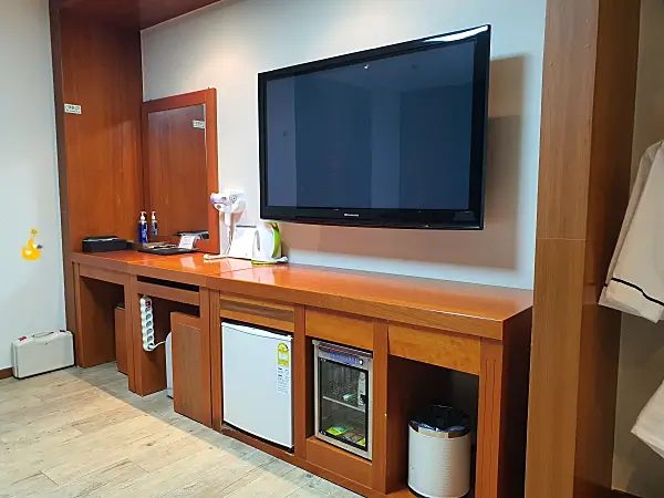 호텔 방에 있는 화장대와 벽에 걸린 TV, 선반 아래 놓인 냉장고와 자외선 소독기가 있는 모습