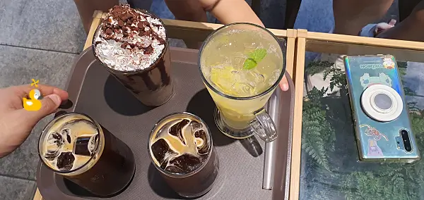 초록색 양치식물이 보이는 투명 테이블 위에 사나고초코라떼, 레몬에이드와 아이스아메리카노가 올려진 쟁반