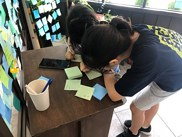 입구에 있는 포스트잇에 펜으로 사나고 카페 방명록을 쓰고 있는 아이들